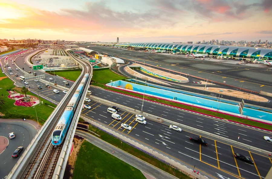 
Aerial view of Dubai International Airport, featuring a tram and highway in the foreground, with terminals visible in the background.